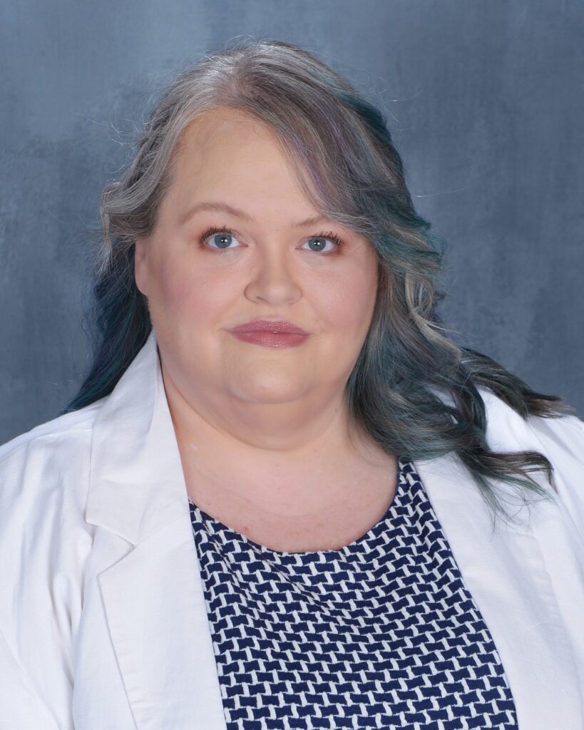 A smiling woman with blue and silver hair wearing a blue dress and white blazer
